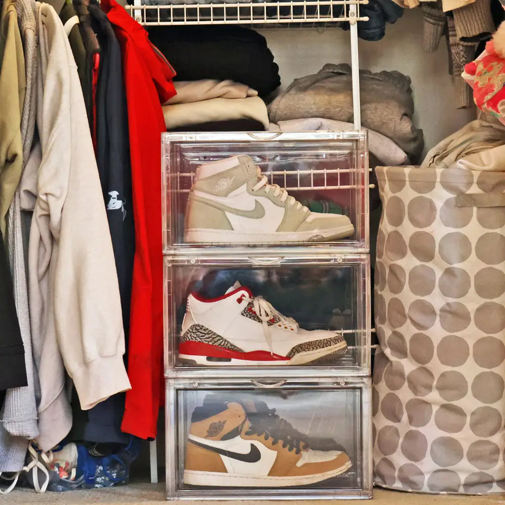 Stack of clear acrylic boxes containing sneakers in a closet with clothes and bags.