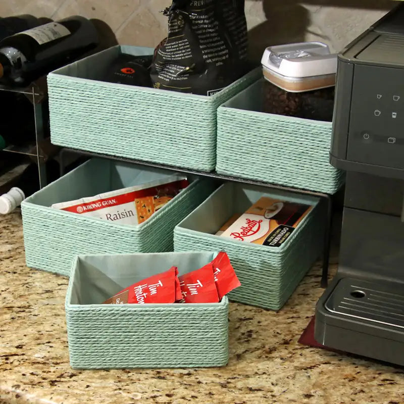Green woven storage bins on a kitchen counter with snacks and coffee items.