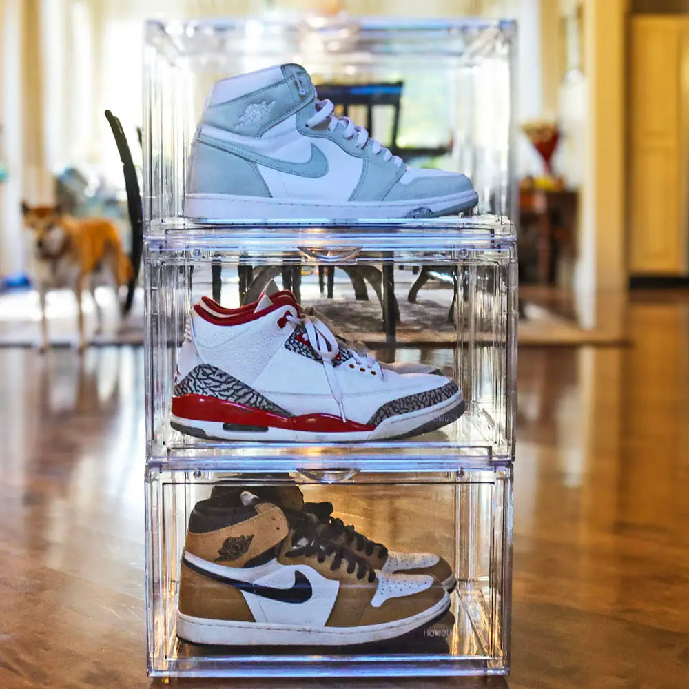 Three pairs of sneakers stacked on clear acrylic boxes in a room with wooden flooring.